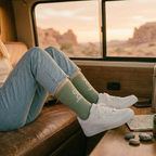 Person sitting inside a vehicle with legs crossed, wearing green socks and white sneakers, with a desert landscape outside.