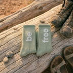 Green socks with 'be free' text on a wooden log with hiking boots.