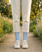 Person wearing light blue socks with 'peace within' text, standing on a path with greenery.
