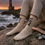 Person wearing beige socks with 'Stay Soft' branding on a rocky beach at sunset.