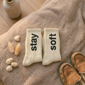 Pair of socks with 'stay soft' text on a textured surface with a vase and slippers in the background.