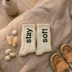 Pair of socks with 'stay soft' text on a textured surface with a vase and slippers in the background.