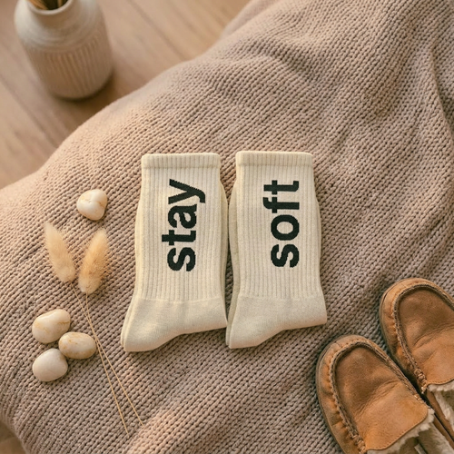 Pair of socks with 'stay soft' text on a textured surface with a vase and slippers in the background.